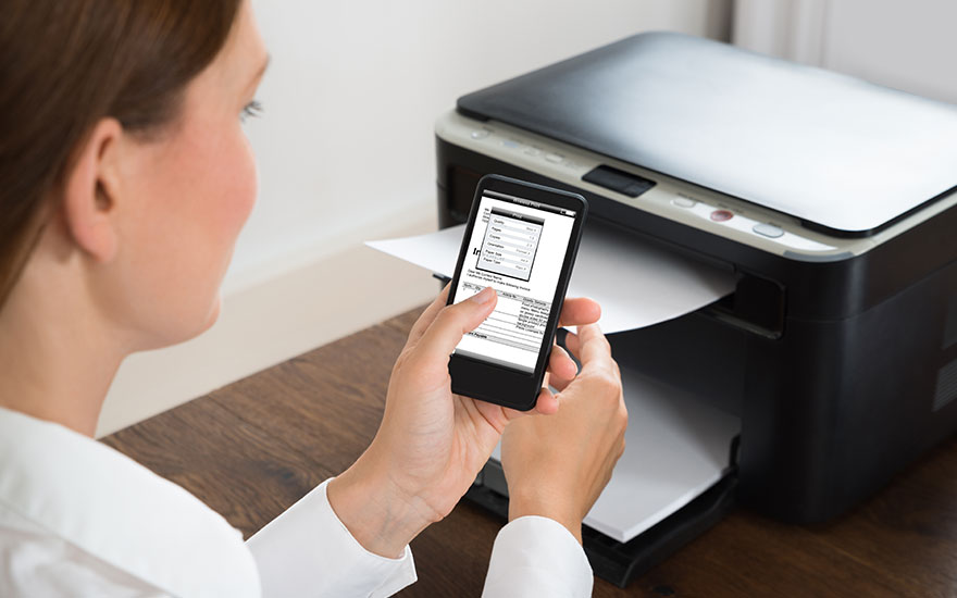 Een printer staat op een houten tafel. Een telefoon wordt voor de printer gehouden. Er komt papier uit de printer.