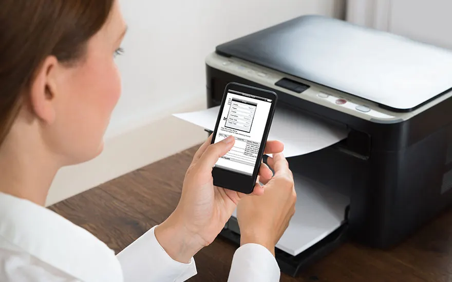 Een printer staat op een houten tafel. Een telefoon wordt voor de printer gehouden. Er komt papier uit de printer.