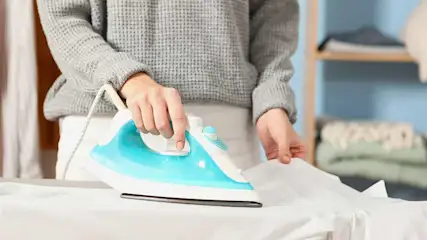 Een blauw-wit strijkijzer strijkt een wit kledingstuk op een strijkplank. Op de achtergrond een rek met opgevouwen kleding.