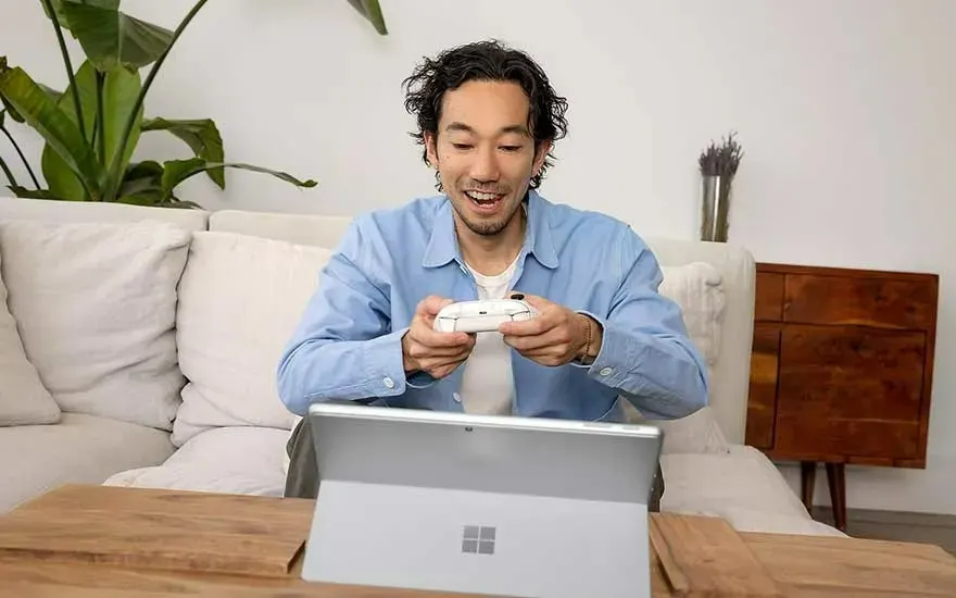 Un ordinateur portable gris est sur une table en bois, avec une manette de jeu blanche. Un canapé et des plantes sont visibles en arrière-plan.