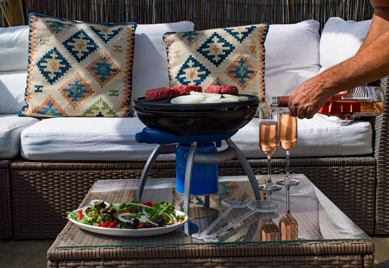 Een buitenscène met een barbecue op een tafel, twee glazen, een salade en kussens op een witte bank.