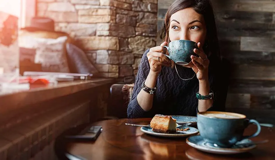 Une table en bois avec une tasse, un gâteau et un téléphone. Une personne boit dans une tasse devant un mur de briques.