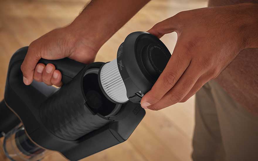 Une personne remplace le filtre blanc d'un aspirateur noir, avec un sol en bois clair en arrière-plan.