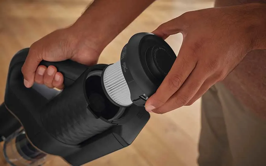 Une personne remplace le filtre blanc d'un aspirateur noir, avec un sol en bois clair en arrière-plan.