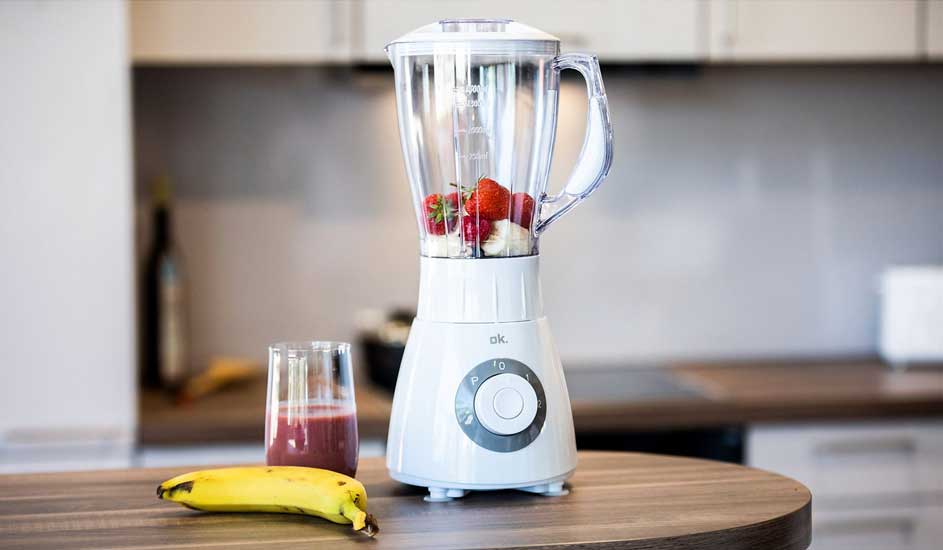Een witte blender met aardbeien en banaan staat op een houten tafel, naast een glas smoothie en een banaan.