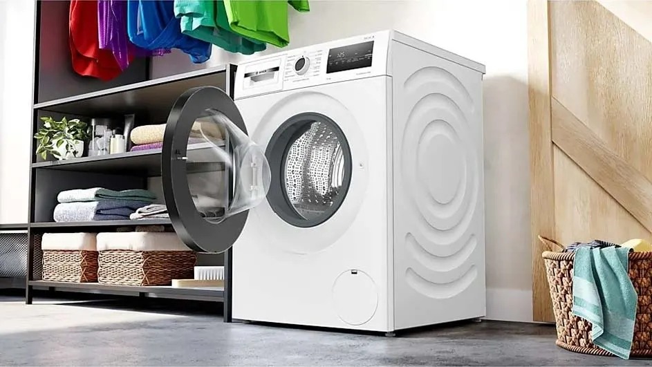 Un lave-linge Bosch est ouvert dans une buanderie moderne, avec des vêtements colorés au-dessus et des paniers, des plantes et des serviettes sur l’étagère.
