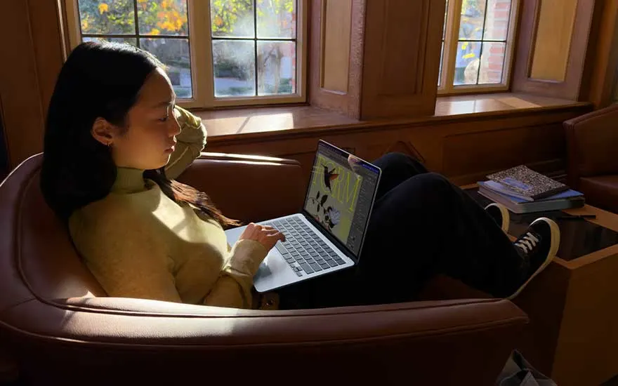 Persoon ontspant in een fauteuil bij een raam, met warme zonnestralen op het gezicht, terwijl zij op een laptop werkt met creatieve inhoud.