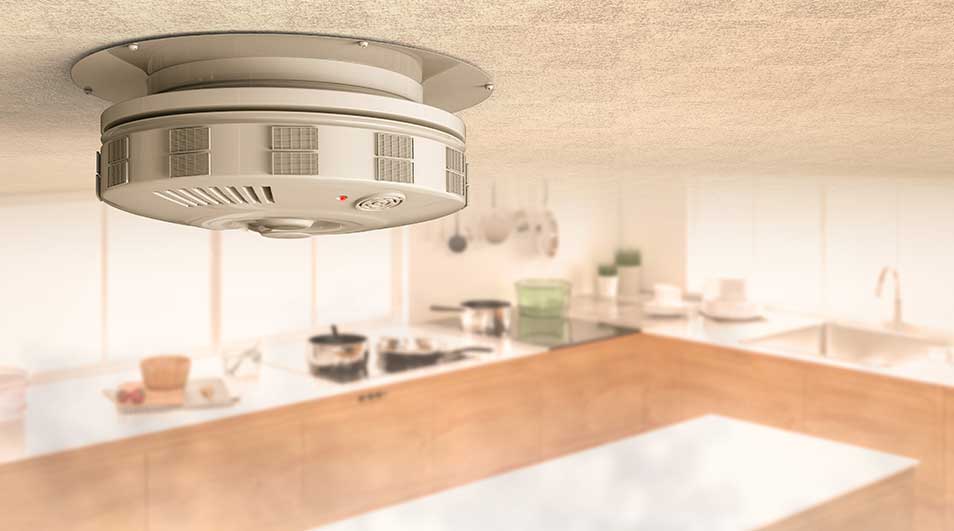 Witte rookmelder aan plafond in keuken. Kookgerei en aanrecht in vervaging op achtergrond.