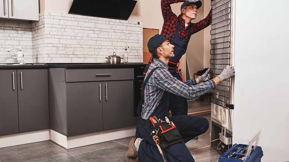 Two repairmen in a kitchen are installing a white refrigerator. Background features gray cabinets and a white brick backsplash.