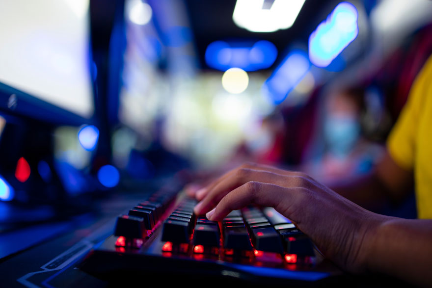 Mains tapant sur un clavier noir rétroéclairé en rouge. Écrans et lumières bleues en arrière-plan. T-shirt jaune visible.