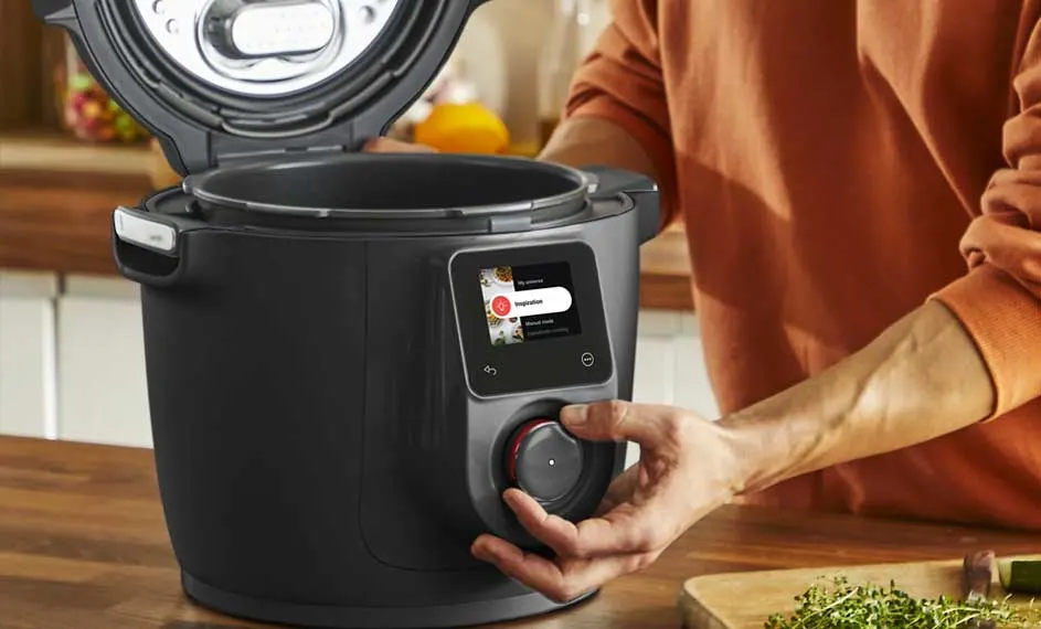 Un autocuiseur noir ouvert, avec un écran tactile et un bouton rotatif, est sur une table en bois dans une cuisine.