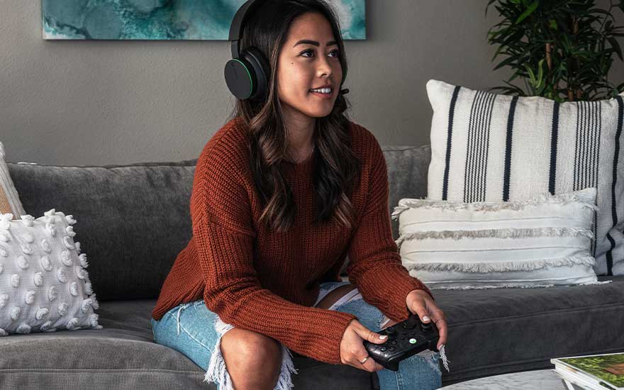 Een vrouw zit ontspannen op de bank met een hoofdtelefoon en controller, terwijl ze geconcentreerd een videogame speelt.