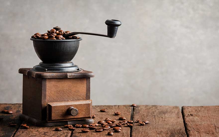 Un moulin à café manuel en bois avec des grains de café sur une table en bois devant un mur gris.