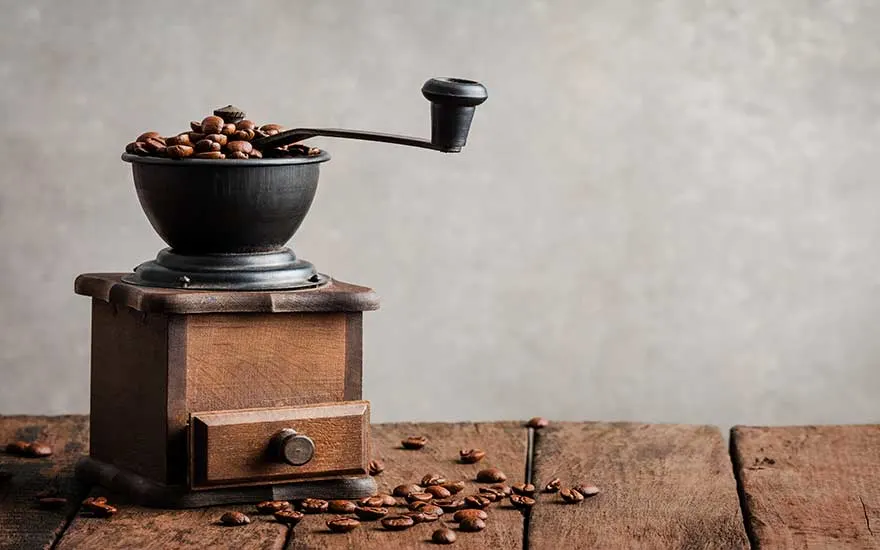 Un moulin à café manuel en bois avec des grains de café sur une table en bois devant un mur gris.