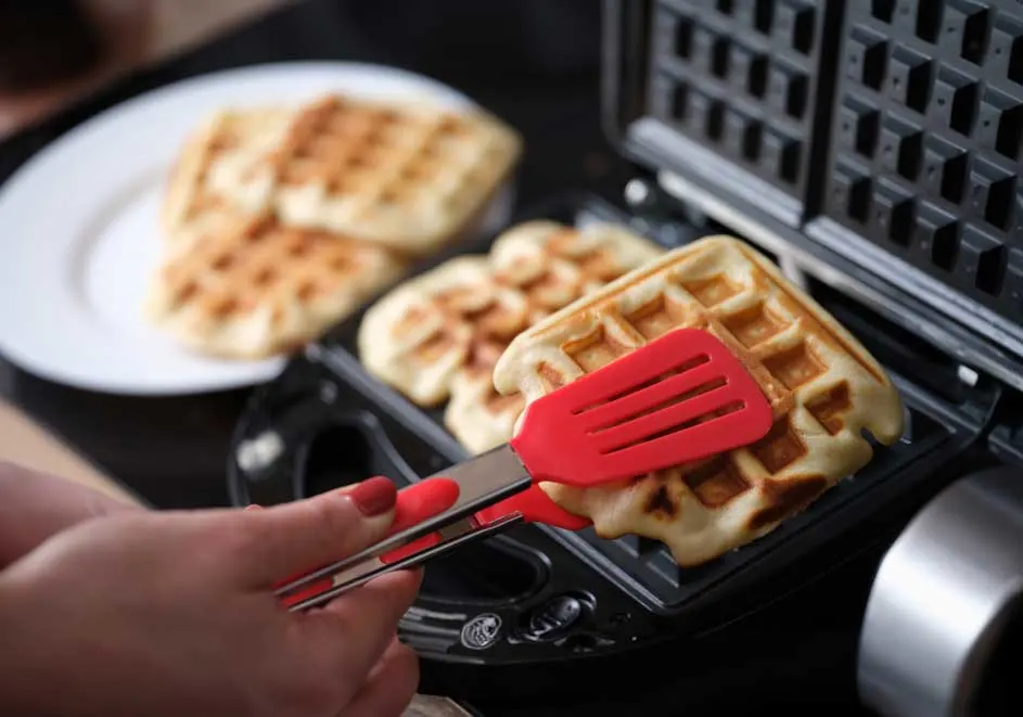 Een wafel wordt uit een zwart wafelijzer gehaald met een rode spatel. Op een wit bord liggen reeds gebakken wafels.