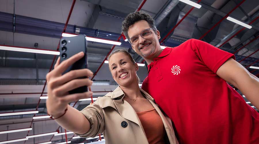 Une femme souriante prend un selfie avec un homme portant un polo rouge dans un environnement commercial moderne.