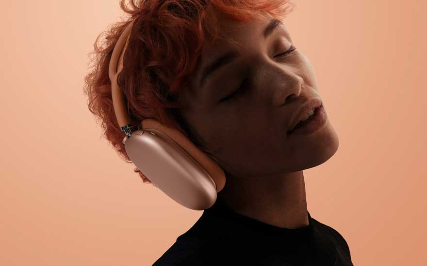 A person with short, coloured hair wears pink over-ear headphones, eyes closed in relaxation against a smooth peach-coloured backdrop.