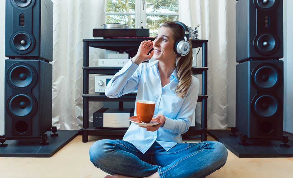 Zittende vrouw met koptelefoon, kopje, en luidsprekers. Apparatuur op een zwarte rek in de achtergrond.