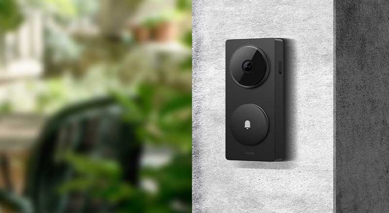 Une sonnette vidéo intelligente noire est fixée sur un mur extérieur, avec un jardin vert flou en arrière-plan.