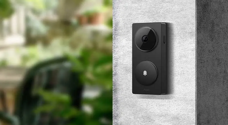 Een zwarte slimme videodeurbel hangt aan een buitenmuur, met een onscherpe groene tuin op de achtergrond.