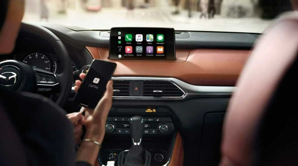 Interieur van een Mazda CX-9 met een scherm met Apple CarPlay en een persoon die een smartphone vasthoudt.