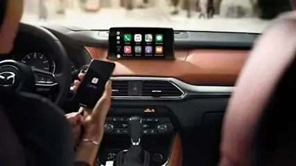 Interieur van een Mazda CX-9 met een scherm met Apple CarPlay en een persoon die een smartphone vasthoudt.