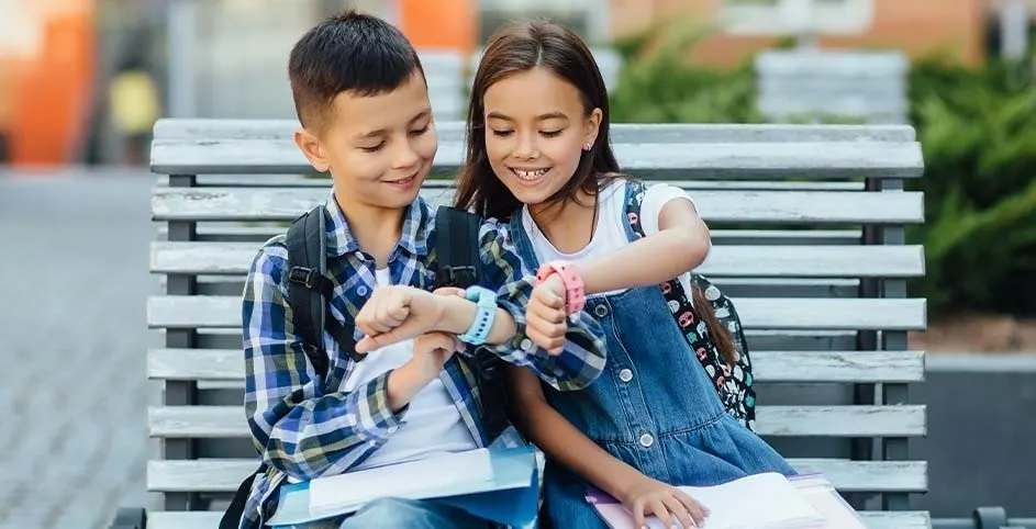 Twee kinderen met rugzakken zitten op een bank en kijken naar hun polshorloges. Achtergrond: gebouw en groen.