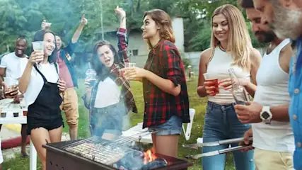 Een barbecue met mensen die drankjes vasthouden. Op de achtergrond staan bomen en een wit hek.