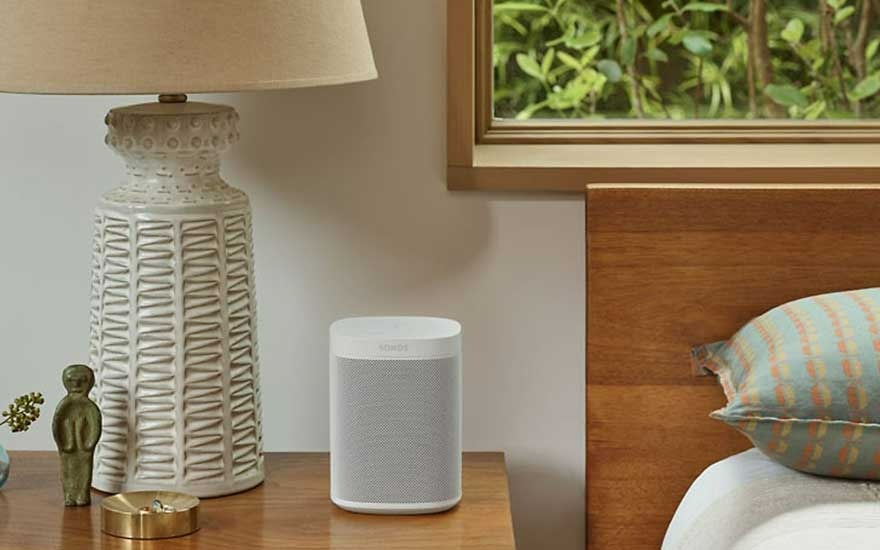 Lampe beige à motif, enceinte grise sur table de chevet en bois. Fenêtre avec verdure visible.