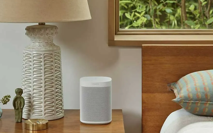 Lampe beige à motif, enceinte grise sur table de chevet en bois. Fenêtre avec verdure visible.