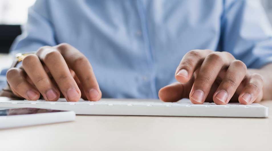 Mains tapant sur un clavier blanc. Un téléphone blanc est visible à gauche. Un homme porte une chemise bleue.