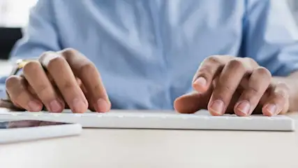 Mains tapant sur un clavier blanc. Un téléphone blanc est visible à gauche. Un homme porte une chemise bleue.