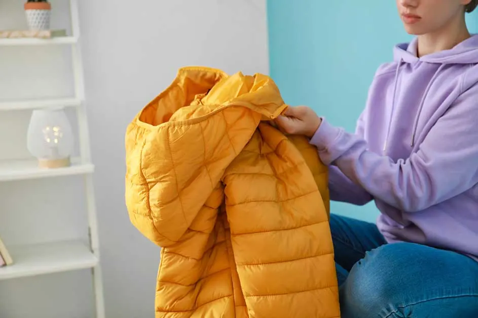 Un manteau jaune matelassé est tenu devant un mur bleu clair. Une étagère blanche est visible en arrière-plan.
