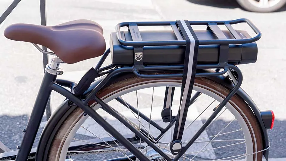 Zicht op een fiets met een bruin zadel en een zwarte batterij vastgemaakt aan een bagagerek met een riem.