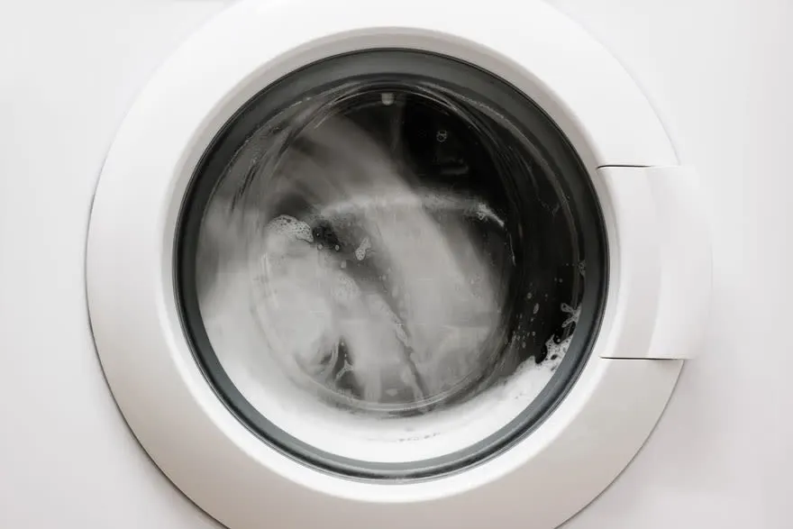Een witte wasmachine met een ronde deur waar kleding in ronddraait met water en zeep.