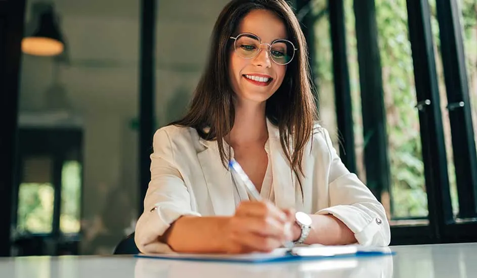 Une personne écrit sur un bloc-notes bleu sur une table blanche. Une fenêtre à barreaux noirs est visible à l'arrière-plan.