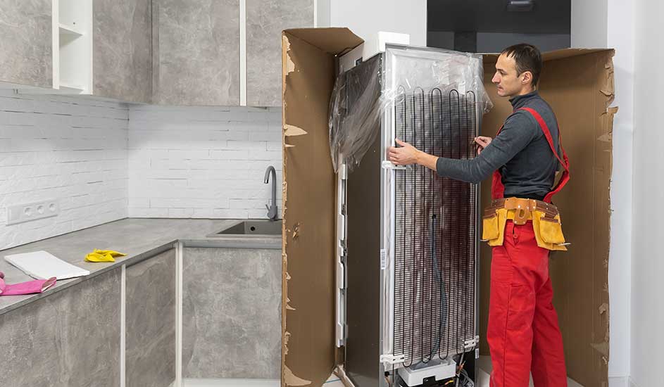 Un frigo neuf, emballé dans du carton, est en cours d'installation dans une cuisine avec des armoires grises.