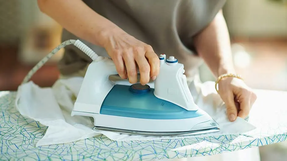 Une personne repasse un vêtement blanc sur une planche à repasser à motifs bleus et verts. Le fer est blanc et bleu.
