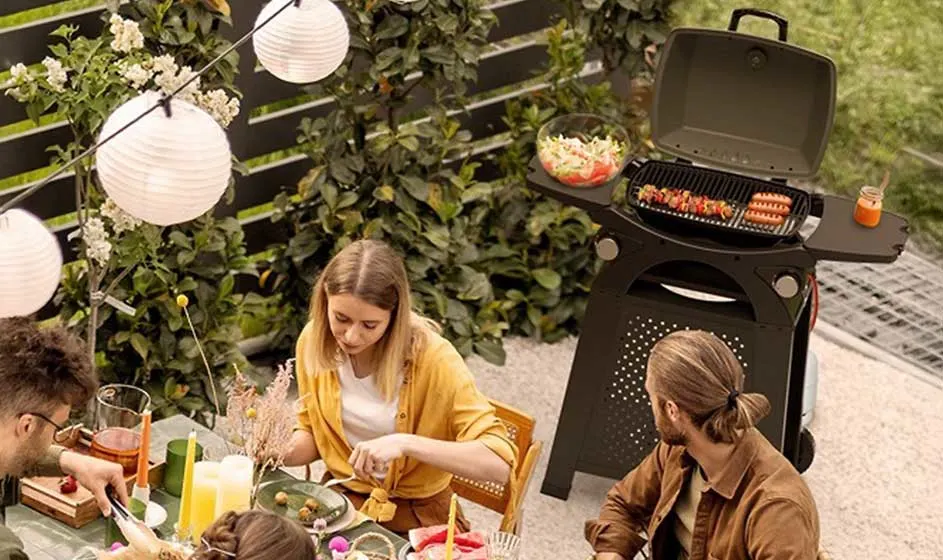 Mensen eten buiten in een tuin, terwijl op een barbecue worstjes grillen tussen lantaarns, planten en zomerlicht.