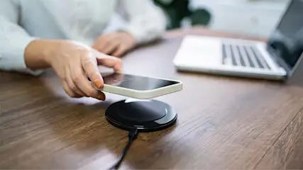 Sur une table en bois, un smartphone est sur le point d'être placé sur un socle de chargement sans fil noir, avec un ordinateur portable en arrière-plan.
