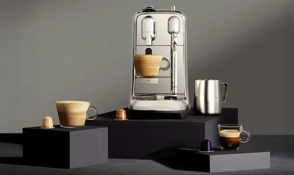 Une élégante machine à café est posée sur des blocs noirs, avec tasses, capsules et pot à lait métallique sur fond gris.