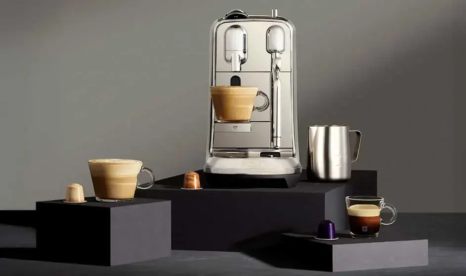 Une élégante machine à café est posée sur des blocs noirs, avec tasses, capsules et pot à lait métallique sur fond gris.