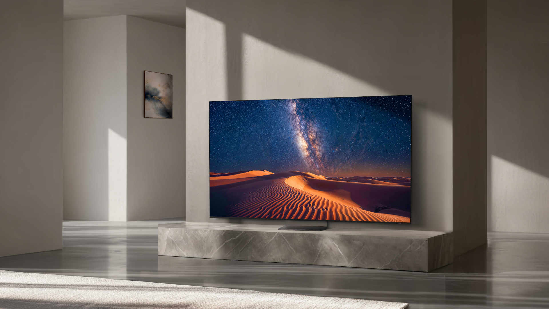 Une grande télévision posée sur un meuble en marbre diffuse une image représentant un paysage désertique et la Voie lactée. Une pièce minimaliste aux murs beiges et baignée de lumière naturelle.