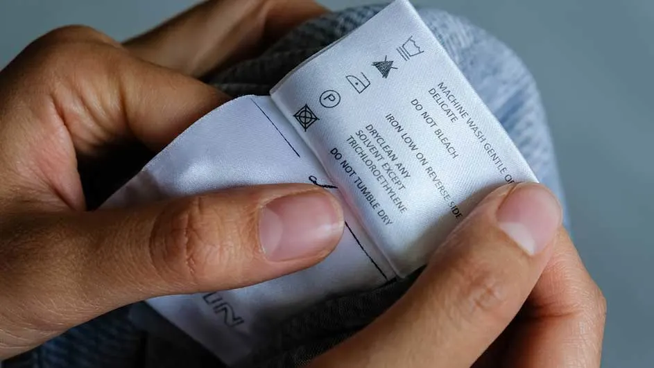 Een hand houdt een wit waslabel vast. Het label toont wasinstructies en symbolen. Grijze stof is zichtbaar op de achtergrond.
