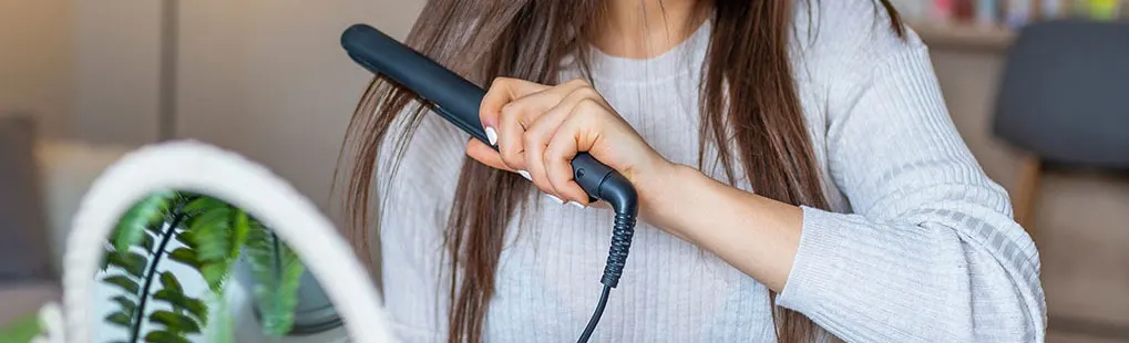 A person is holding a black hair straightener to style brown hair. A ring light and plants are visible in the foreground.