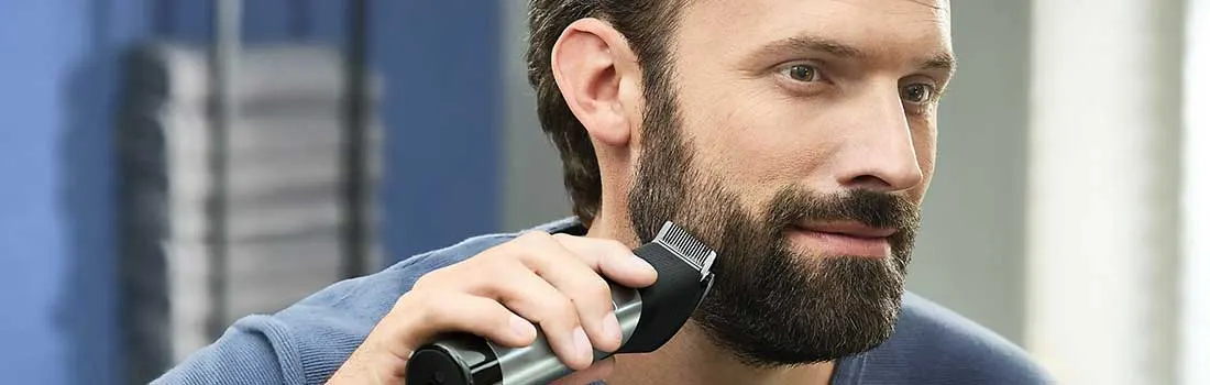 A man uses a black and silver electric trimmer on his beard, in front of a blue and gray background.