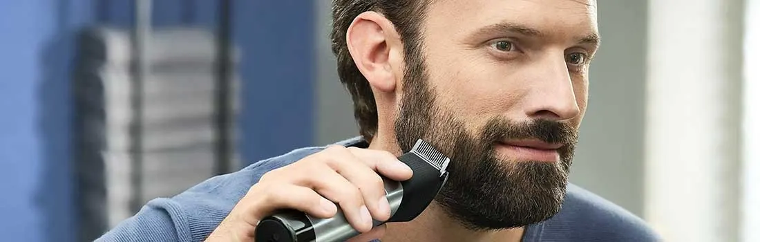 Un homme en t-shirt bleu taille sa barbe avec une tondeuse électrique dans une salle de bain moderne bleue.