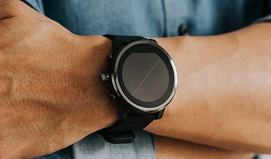 Une smartwatch noire à cadran rond est portée au poignet ; la personne porte une chemise bleu-gris en arrière-plan.
