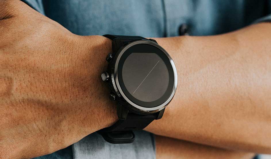 Een zwarte smartwatch met ronde wijzerplaat wordt om een pols gedragen; de drager draagt een blauwgrijs hemd op de achtergrond.