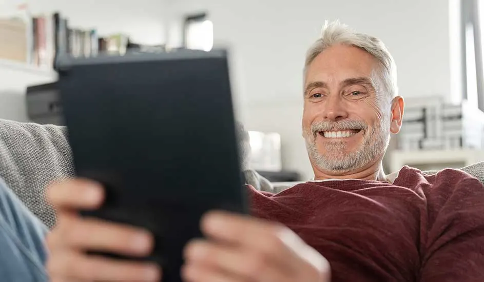 Une personne est assise confortablement sur un canapé dans un salon lumineux, tenant une tablette pour lire ou naviguer.